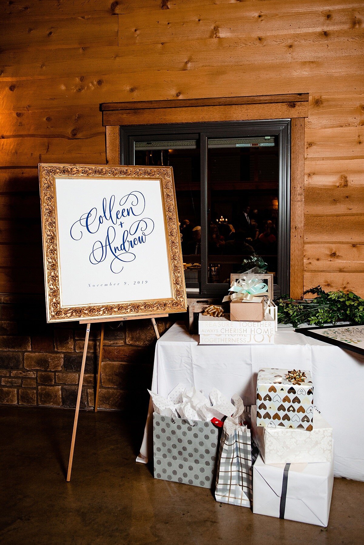 Wedding day gift table overflowing with gifts and gift bags next to a gold framed sign with the bride and groom's names