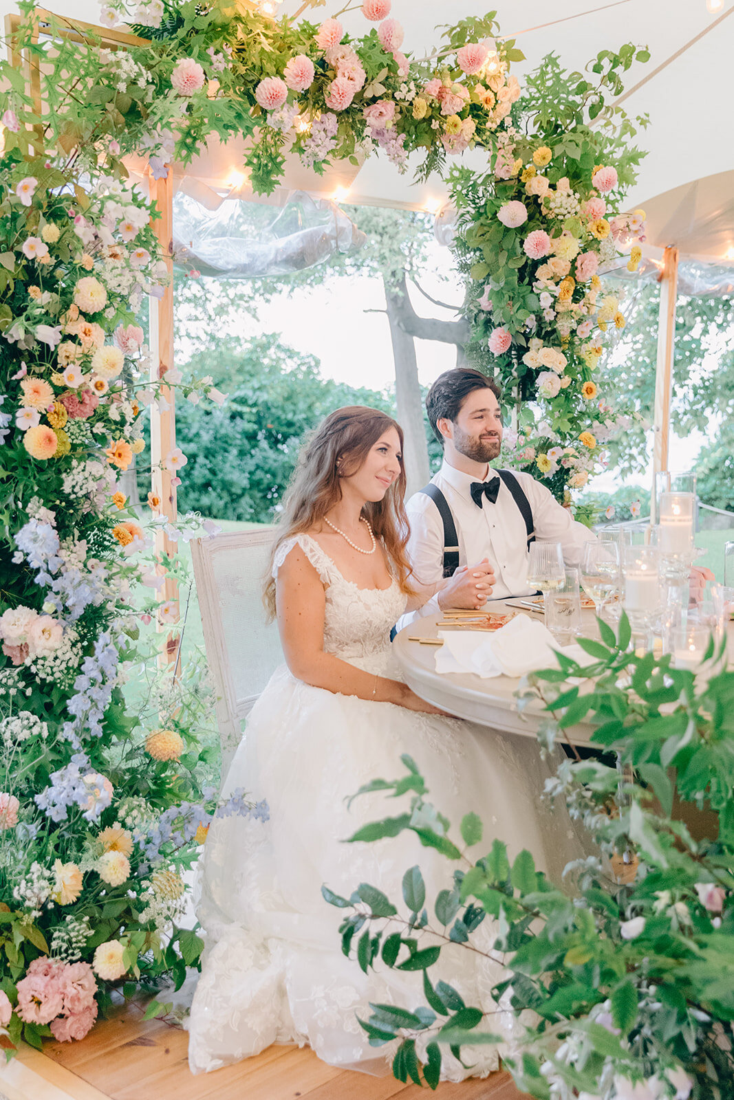 A couple sit surrounded by an arch of bright pastel flowers at their Montreal wedding reception. 