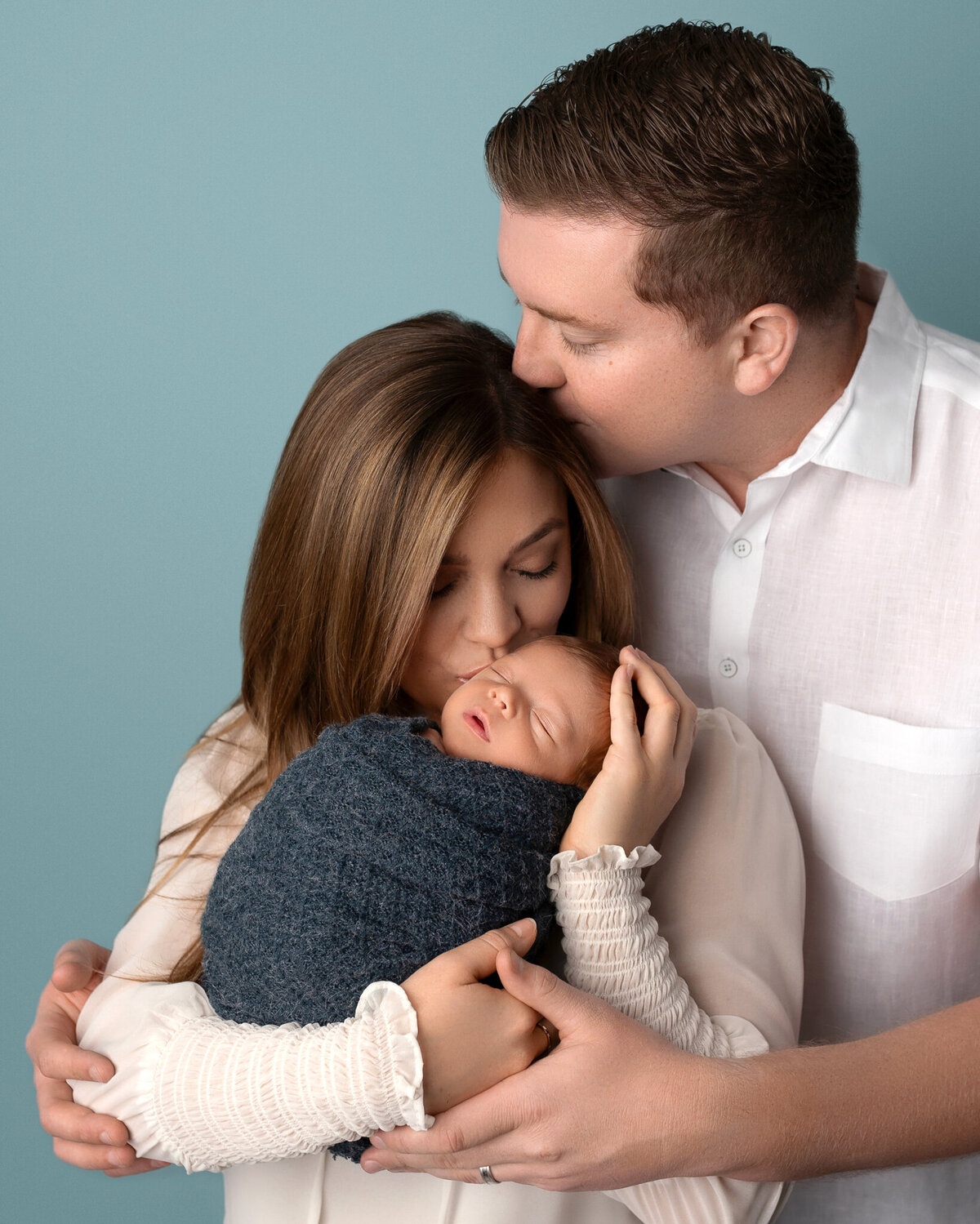 Newborn boy swaddled in blue, held lovingly by parents against soft blue backdrop – Austin family newborn photographer