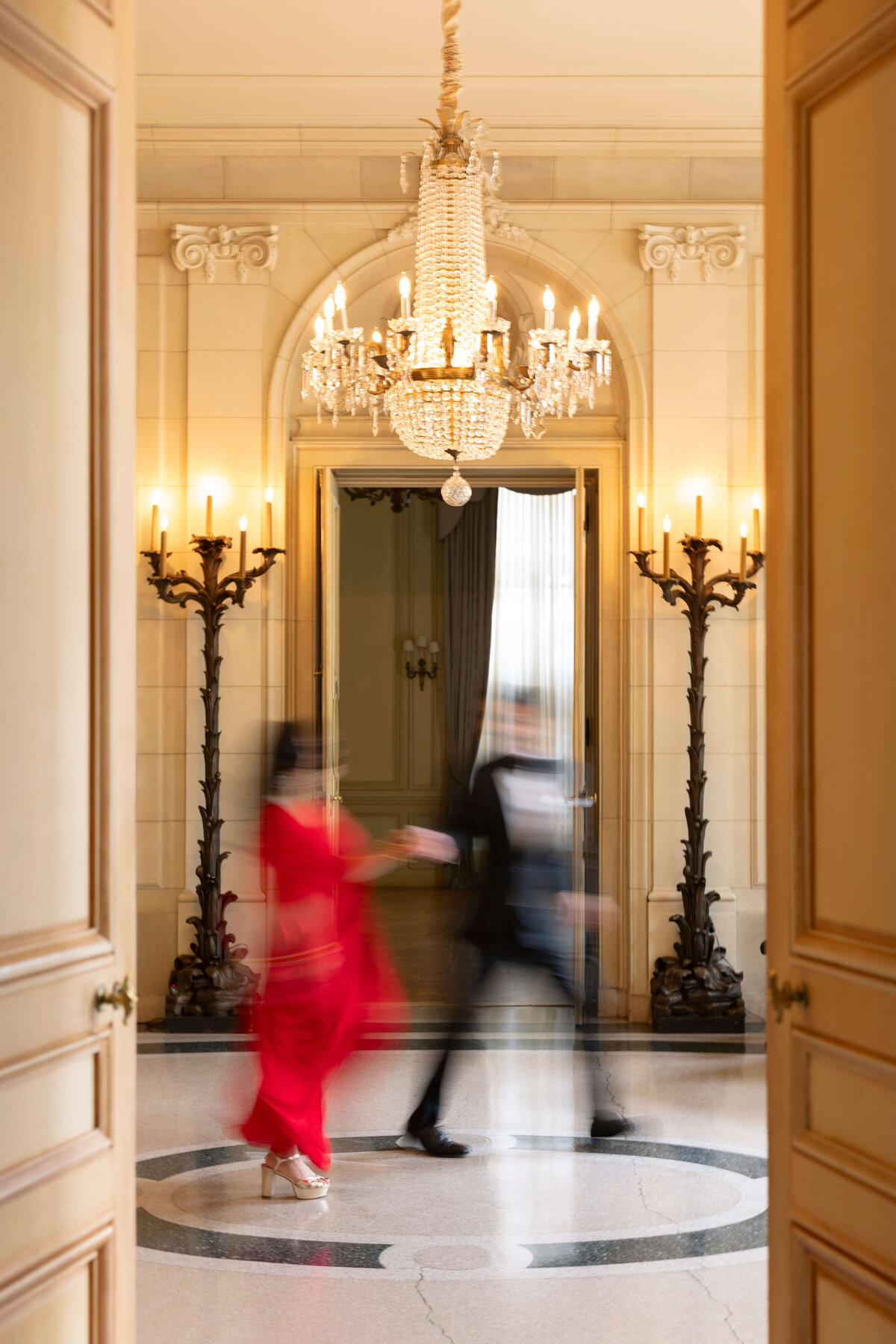A blurred motion image of a person in a red outfit and another in a black suit walking hand in hand through an elegant hallway. The room features a grand chandelier, ornate columns, and double wooden doors, creating a sense of luxury and movement.