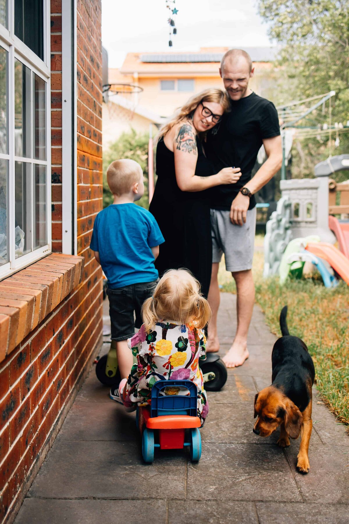 Man and woman with arms around each other, in their backyard, with son, daughter and dog in foreground. Melbourne Photography Gift Vouchers 