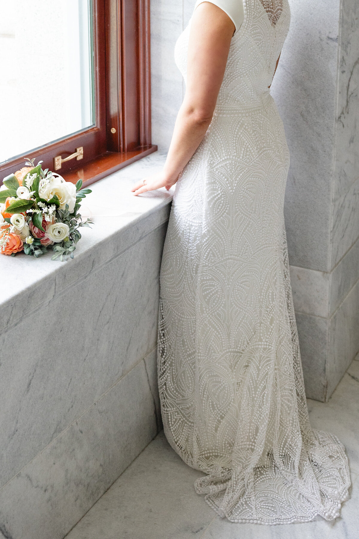 A close-up of a bride’s intricately beaded lace gown and her bouquet of white and orange flowers, resting on a marble windowsill.