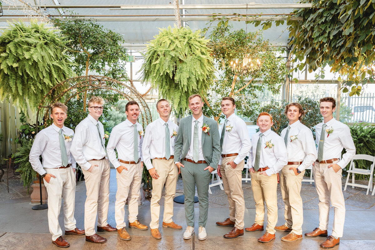 The groom stands at the center of his groomsmen, all dressed in white shirts, beige pants, and sage green ties, posing for a group photo in the wedding venue.