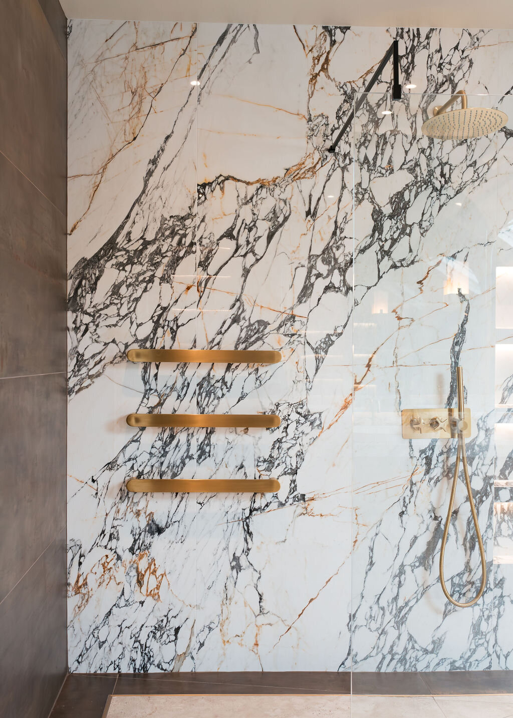 A luxurious shower features a marble wall with black and gold veining. Three gold towel bars are mounted on the left side of the wall. A gold rainfall showerhead and a handheld shower attachment are on the right, with modern fixtures and controls.