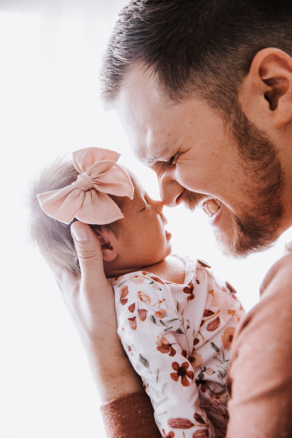father with a beard holds his newborn baby girl and touches his nose to hers as he smiles big