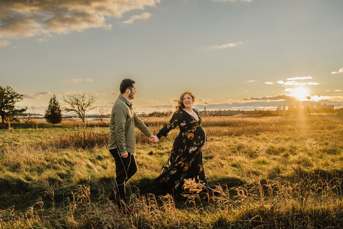 pregnant couple walk hand in hand through a field at sunset