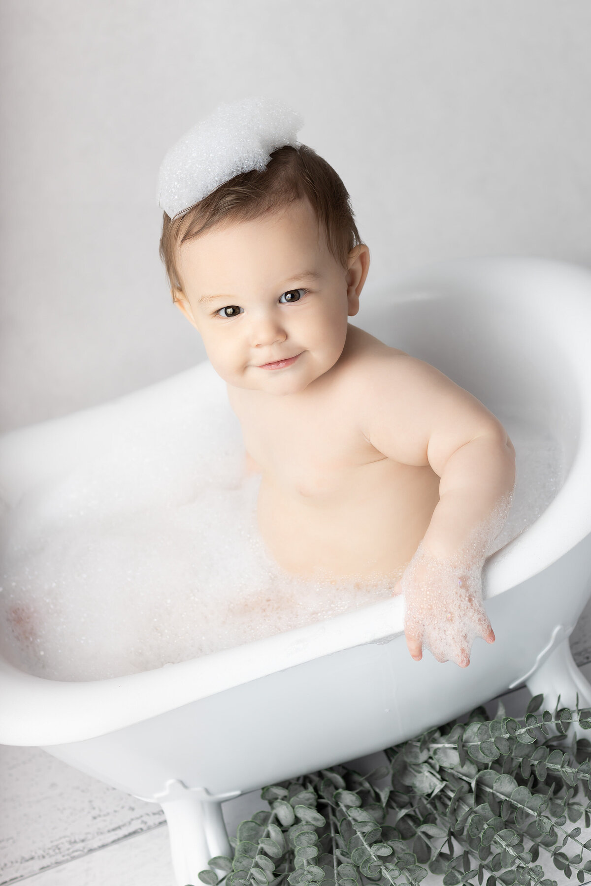 Dallas milestone photographer captures a baby smiling in a white clawfoot tub with a crown of bubbles on their head during a bubble bath session.