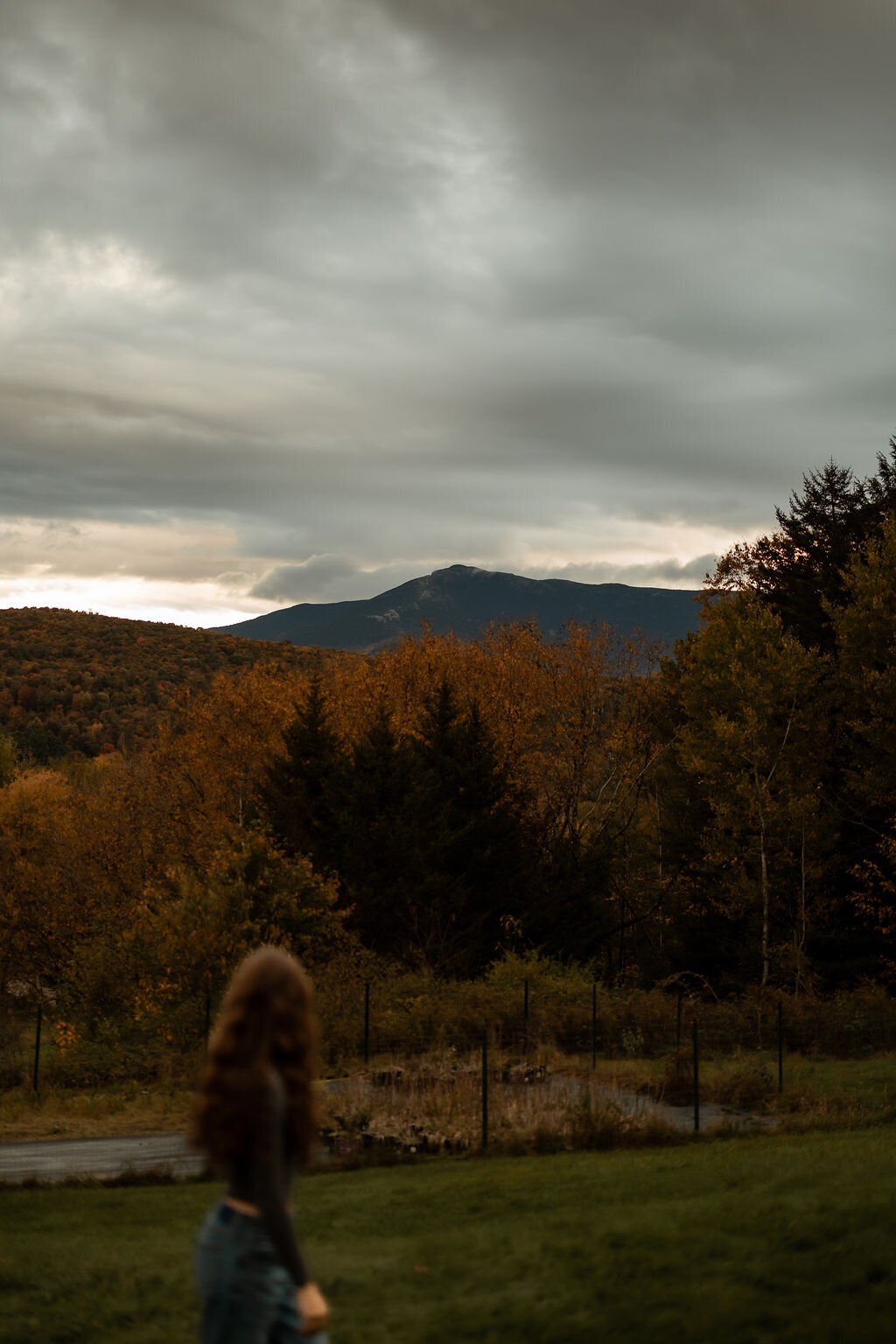 A person with long hair stands on a grassy hill, gazing at distant mountains under a cloudy sky. The surrounding trees show off their autumn colors, crafting a serene scene perfect for New England portrait photography.