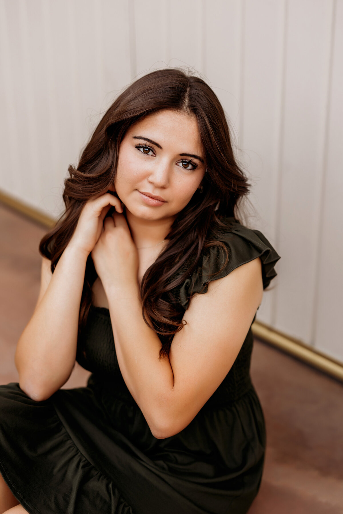 High school senior girl in an olive green dress poses with her hands on her cheek in front of white barn doors for her senior session