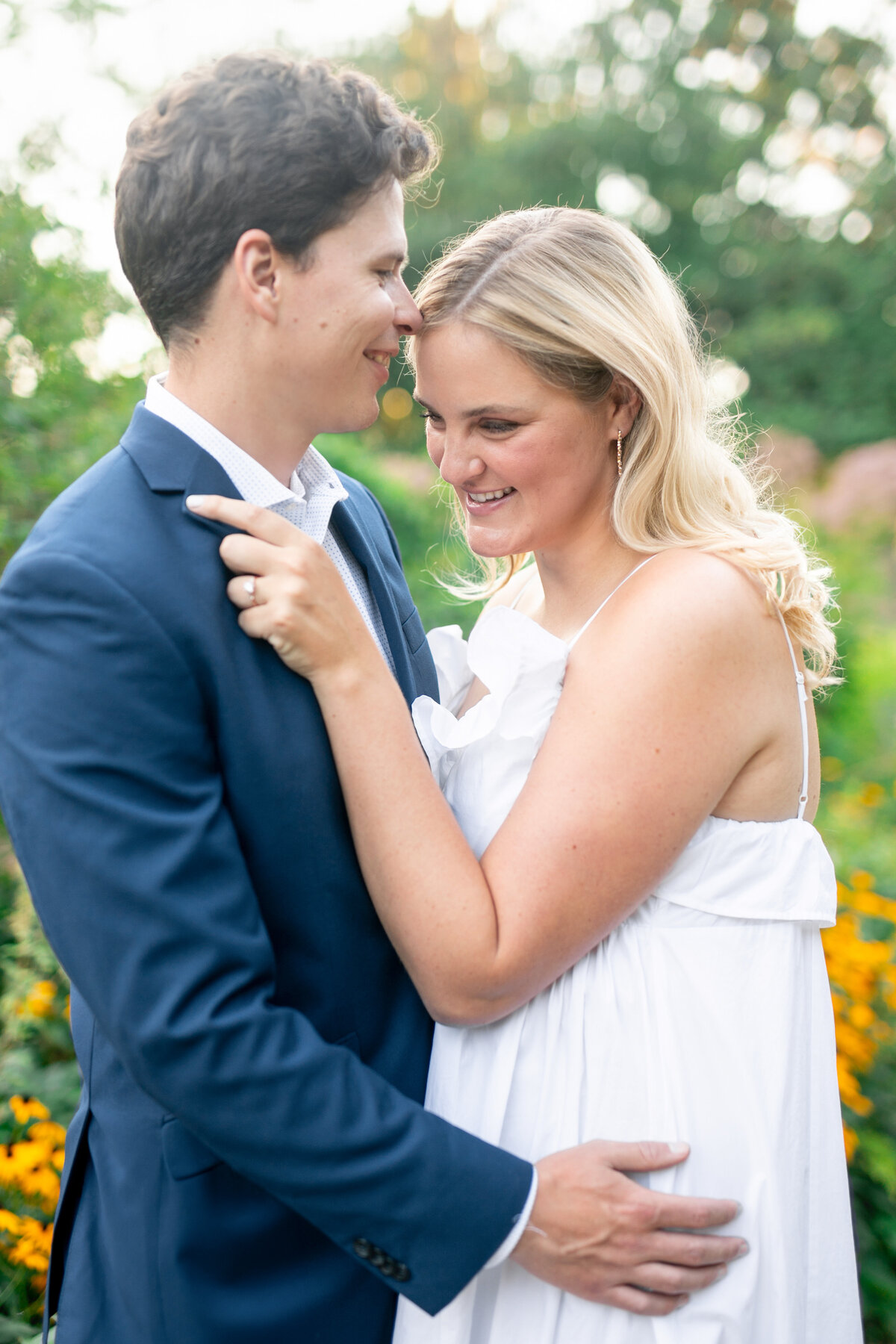 A couple embraces in a garden, with greenery and yellow flowers in the background. The man, wearing a blue suit, leans in toward the smiling woman, dressed in a white sleeveless dress. The setting is bright and cheerful.