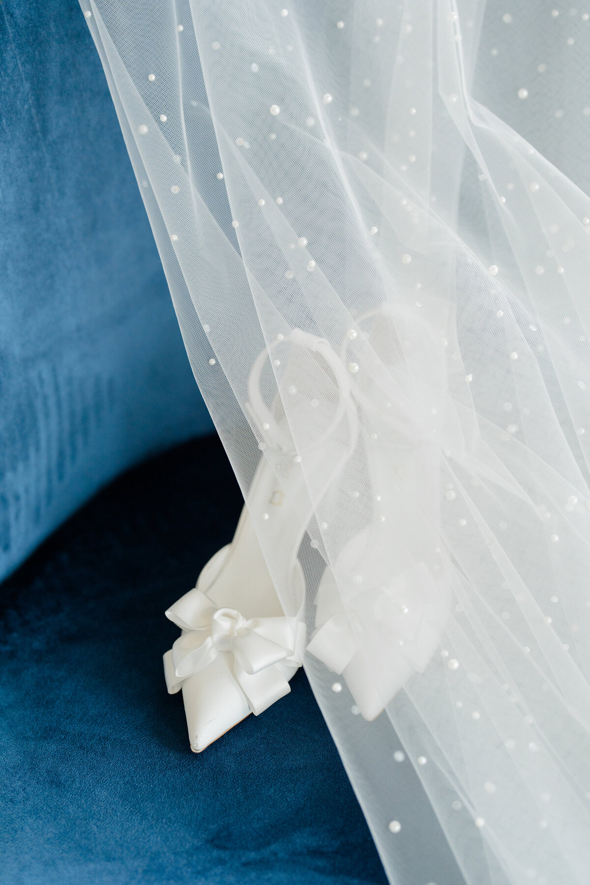 wedding heels with bows sitting on a dark blue suede chair with a pearl detailed veil