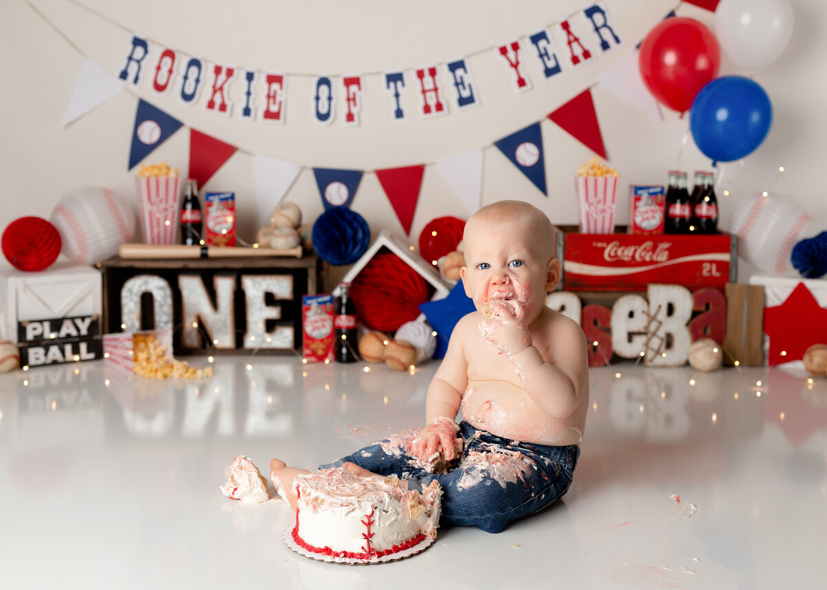 Baseball themed cake smash at West Palm Beach and Boca Raton, FL cake smash photographer. Baby boy wearing blue jeans is eating a baseball bake. His jeans are covered in cake. In teh background, there is red white and blue baseball decor and a "Rookie of the Year" sign.