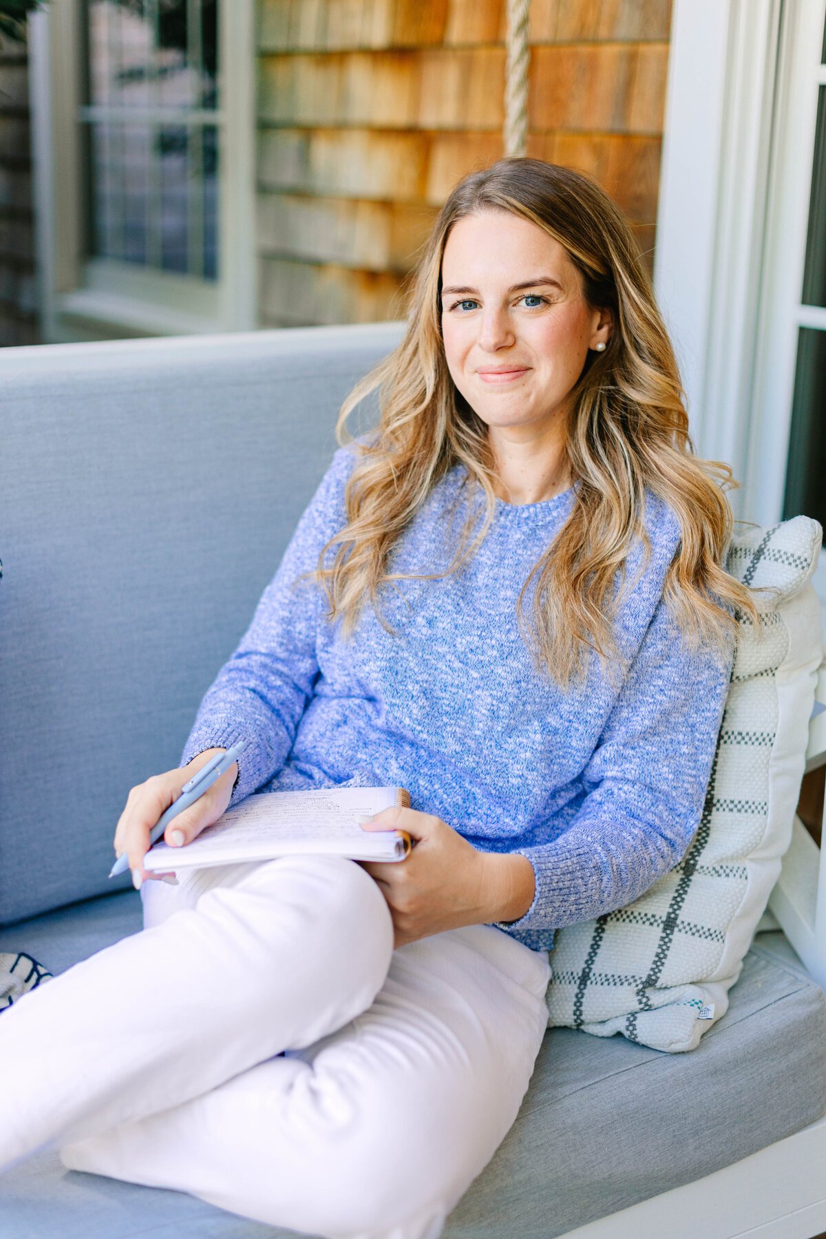 A woman with wavy blonde hair sits on a cushioned porch swing, wearing a cozy blue sweater and white pants. She holds a pen and notebook, with a soft smile on her face. The background features a shingled house exterior and a plaid pillow.