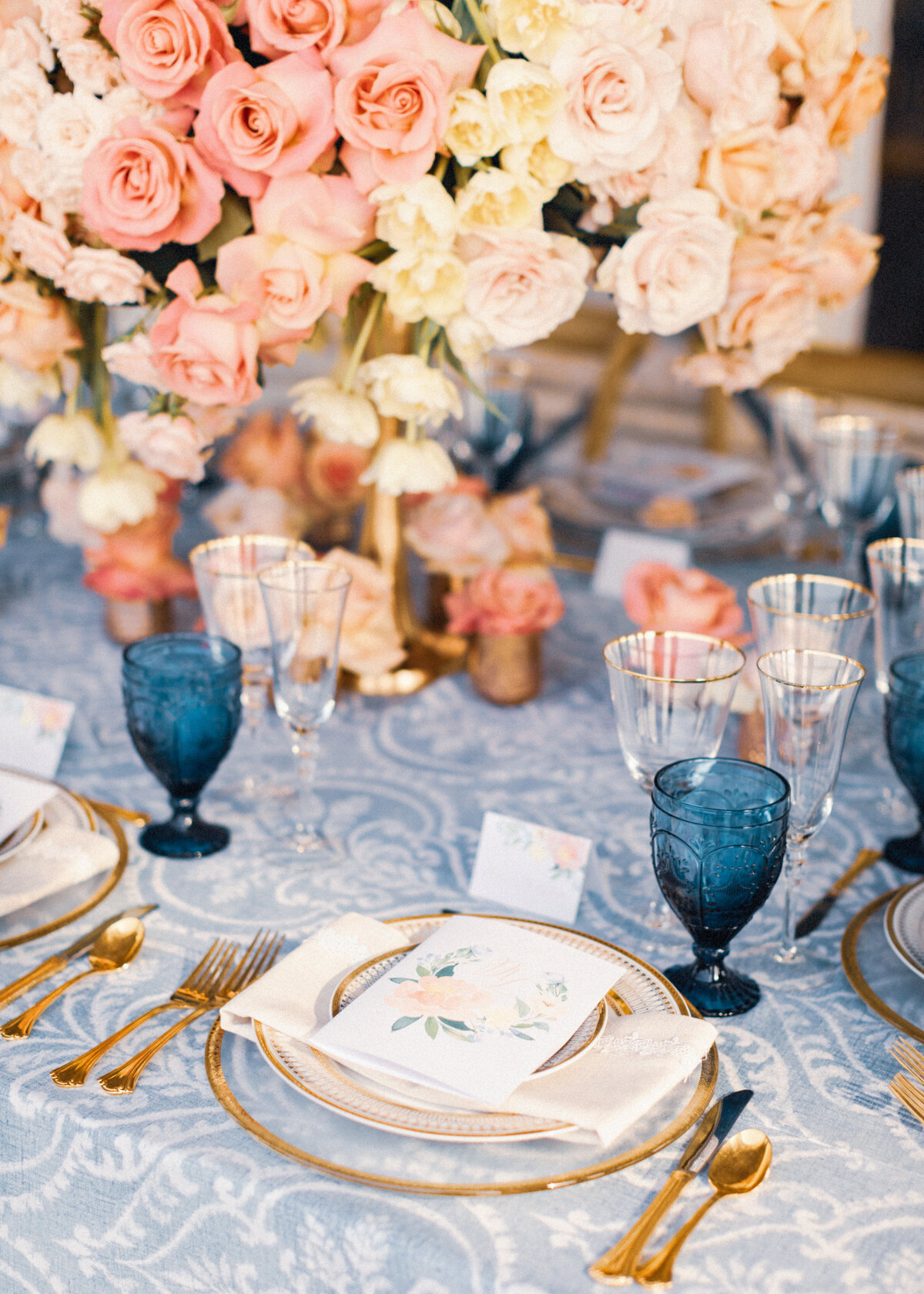 A wedding table is set with vintage lace, blue glassware, gold plates and cutlery and floral menus at a luxury wedding.