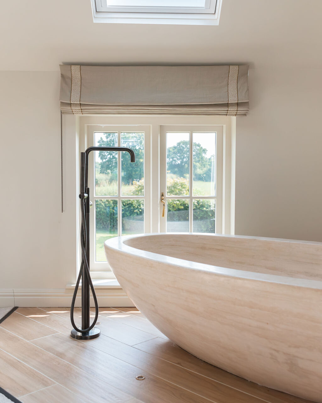 A minimalist neutral bedroom features a large, freestanding wooden bathtub under a skylight. There's a floor-mounted faucet beside the tub next to a window. The space is bright with natural light and neutral tones.