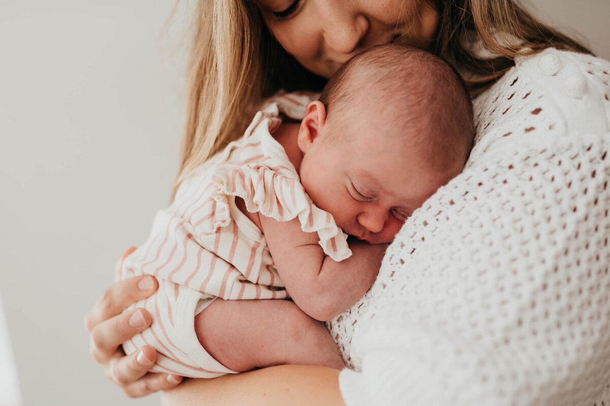 A mother wearing a white sweater holds her sleeping baby against her chest as she gives her baby a soft kiss on the head