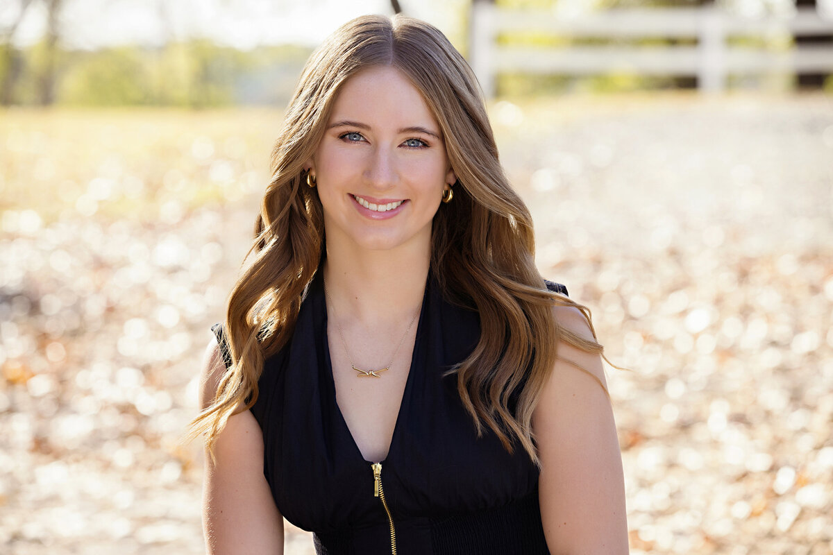 Seated high school senior close-up portrait in black dress with golden leaves on ground and white fence blurry in background
