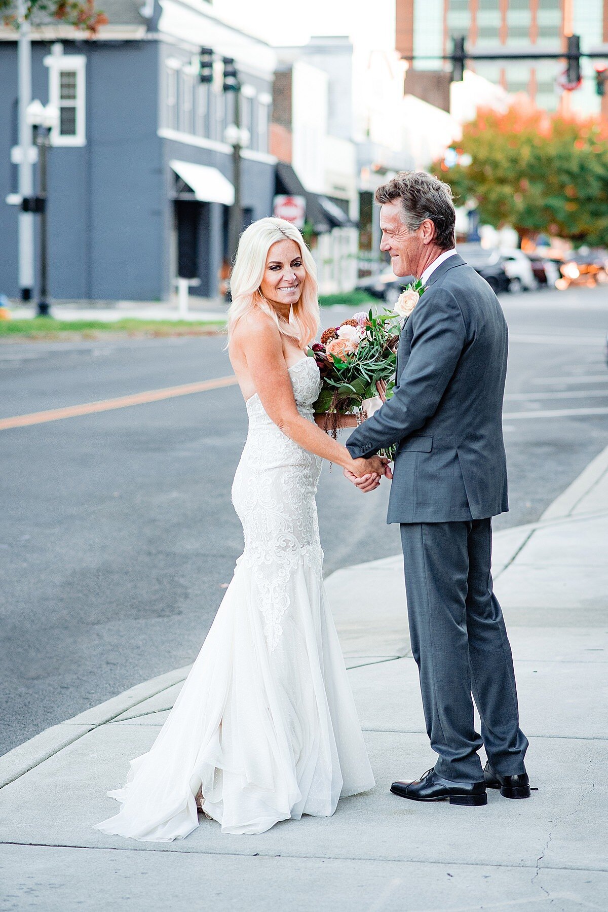 The bride and groom walk down the streets of Murfreesboro. A dark gray building an d office tower are in the background. The bride is wearing a mermaid style dress with a flowing skirt and sweep train. She is smiling at the camera. The groom holds the bride's hand and looks over at her. He is wearing a charcoal gray suit with a blush boutonniere. The bride is carrying a large bouquet of peach, burgundy, blush and ivory flowers with greenery.