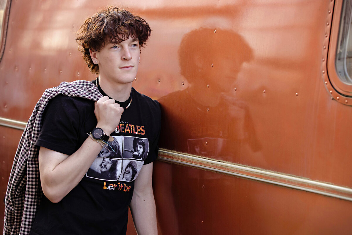High school senior male wearing a classic Beatles t-shirt with a checkered jacket draped over his shoulder, leaning casually against an vintage orange trailer in Charlotte NC with his reflection subtly visible in the metal surface.