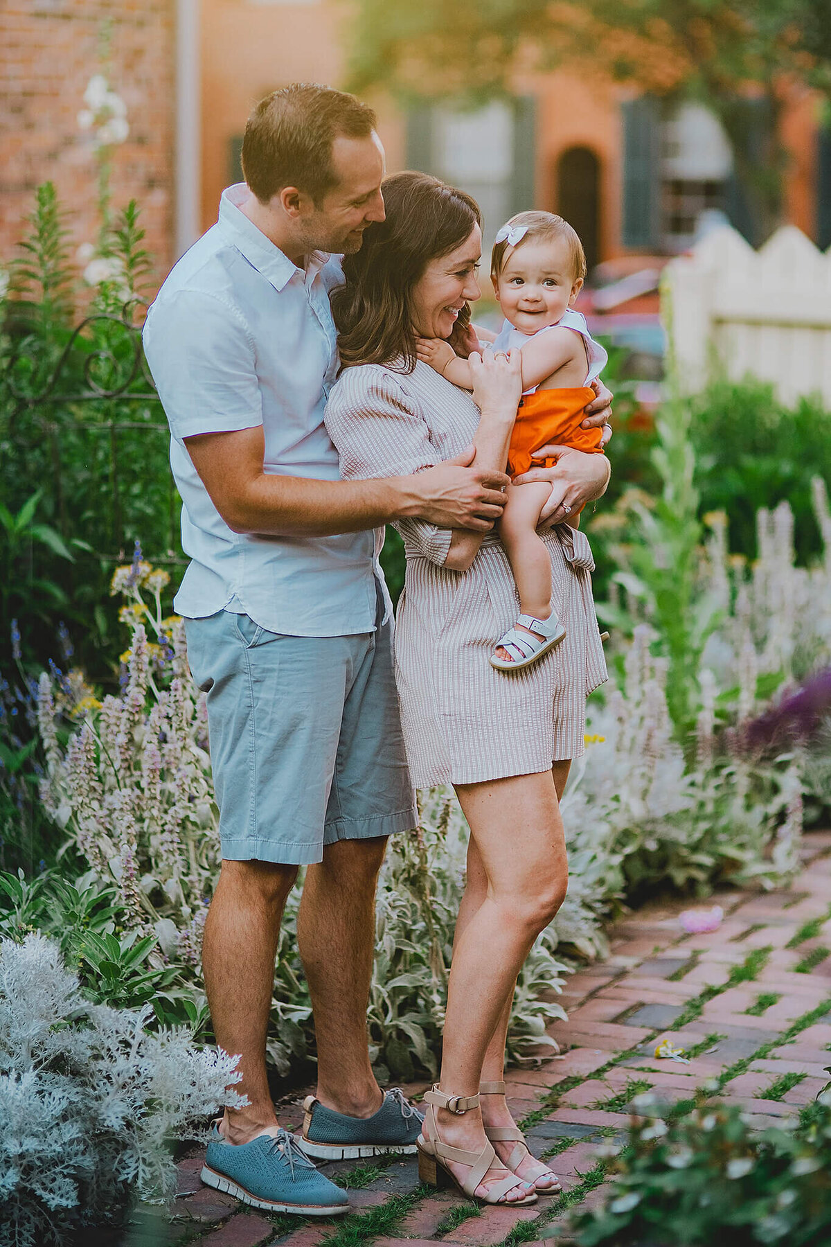 Mother and father smiling and hugging a baby girl in orange shorts in a garden in Fells Point Baltimore Maryland
