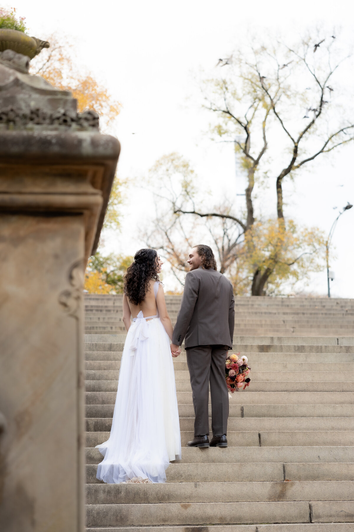 Bride and groom walking up the steps in Central Park, NYC, as birds fly around them during their fall elopement.