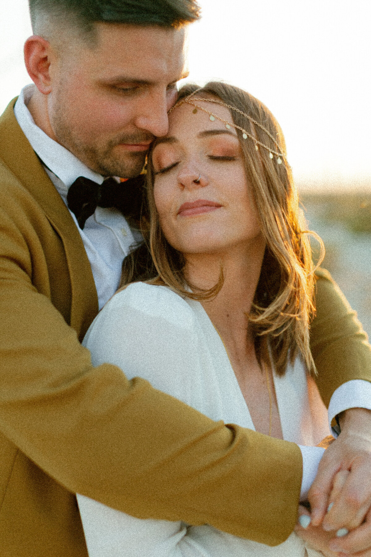 a bride leaning back into her husband as he wraps her arms around her and snuggles into the side of her face as they go in for a warm embrace.