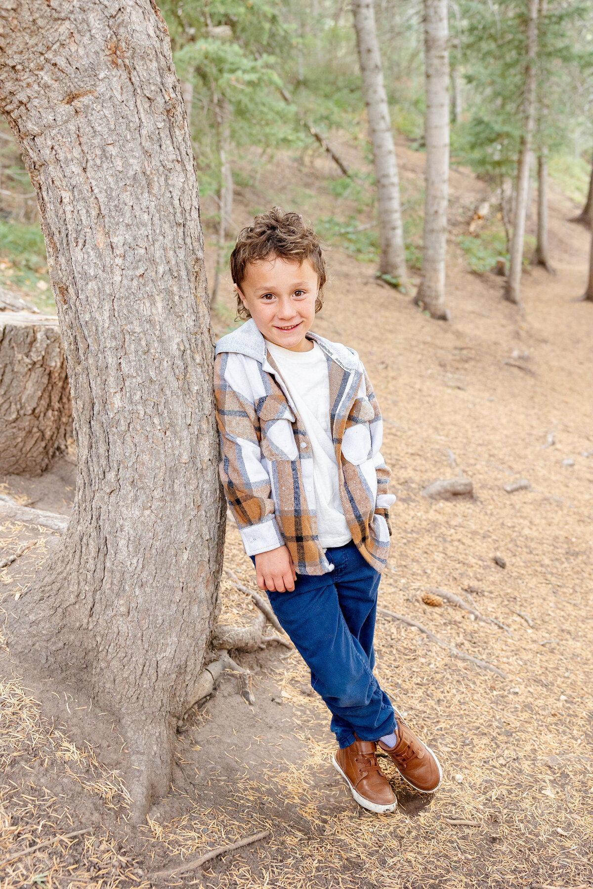 A young boy with curly brown hair leans against a tree in the woods, wearing a plaid flannel jacket, blue pants, and brown shoes, smiling at the camera.