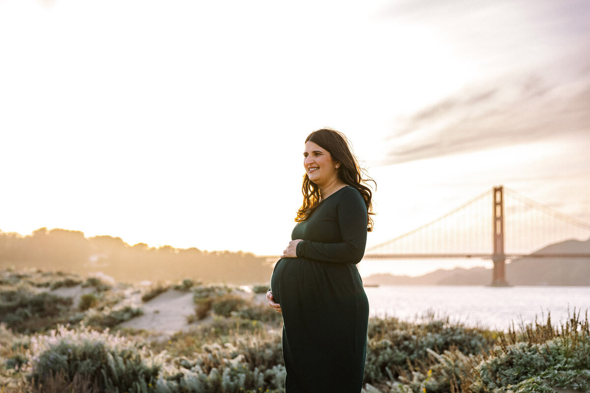 pregnant woman in dark green dress holding baby bump at beach looking away with golden gate bridge in san francisco in the background