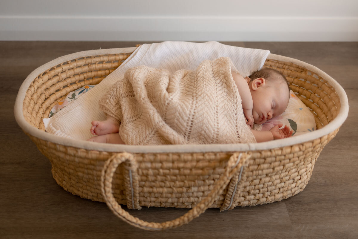 A newborn baby girl is photographed asleep in a rattan bassinet.