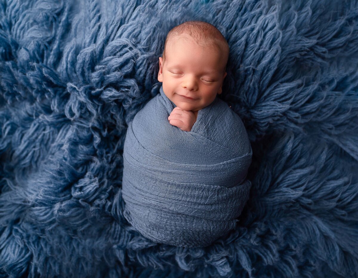 Sleeping baby boy swaddled in blue has his hands peeking out of the swaddle. His eyes are closed but smiling. Baby is laying atop of a blue furry rug. Captured by Main Line photographer Liza Nicole.