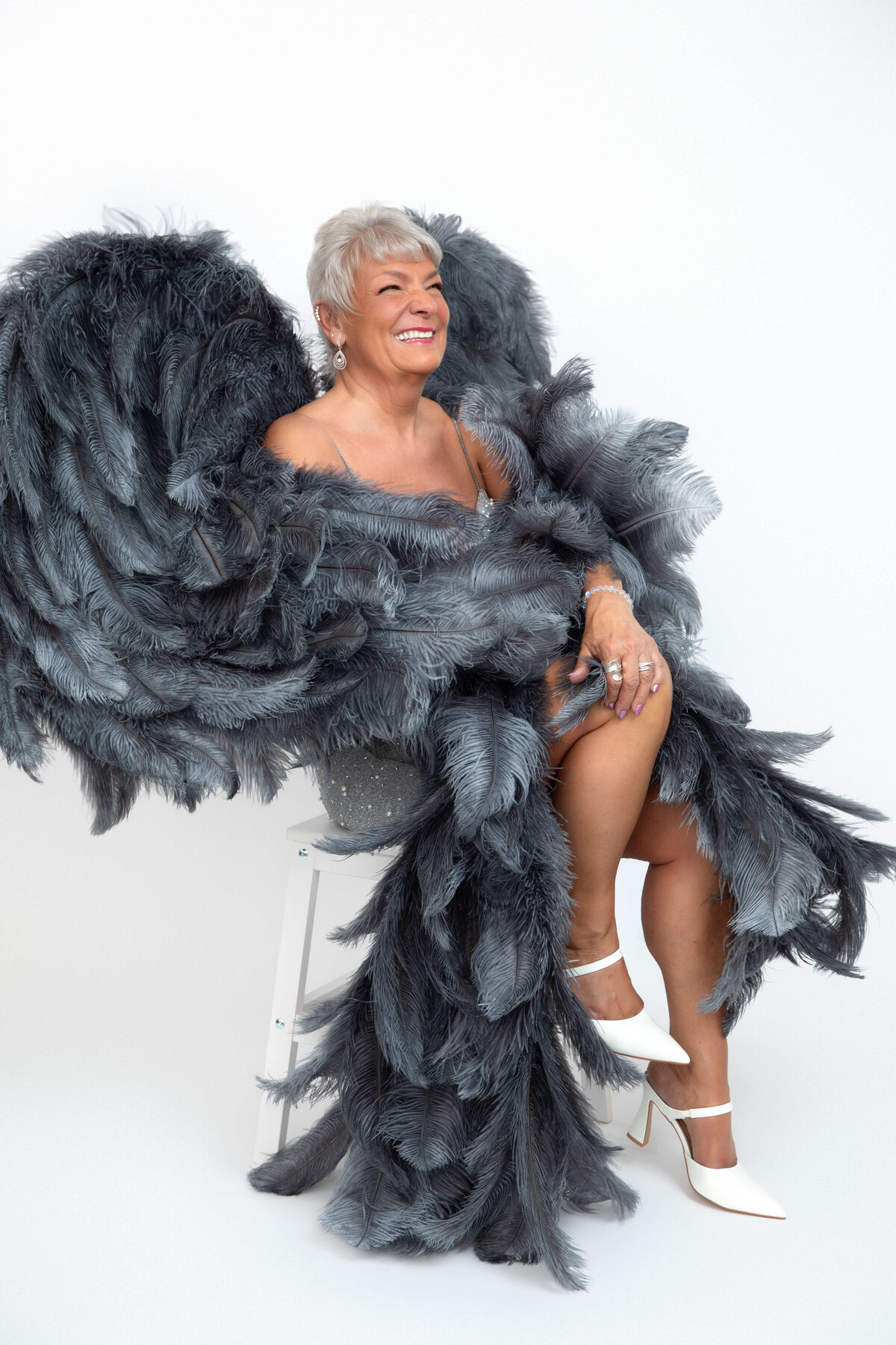 A smiling elderly woman with short white hair is sitting on a white stool, wearing an extravagant feathered garment with large dark feathers that create a wing-like effect. She has white high heels and a glamorous pose against a simple white backdrop.