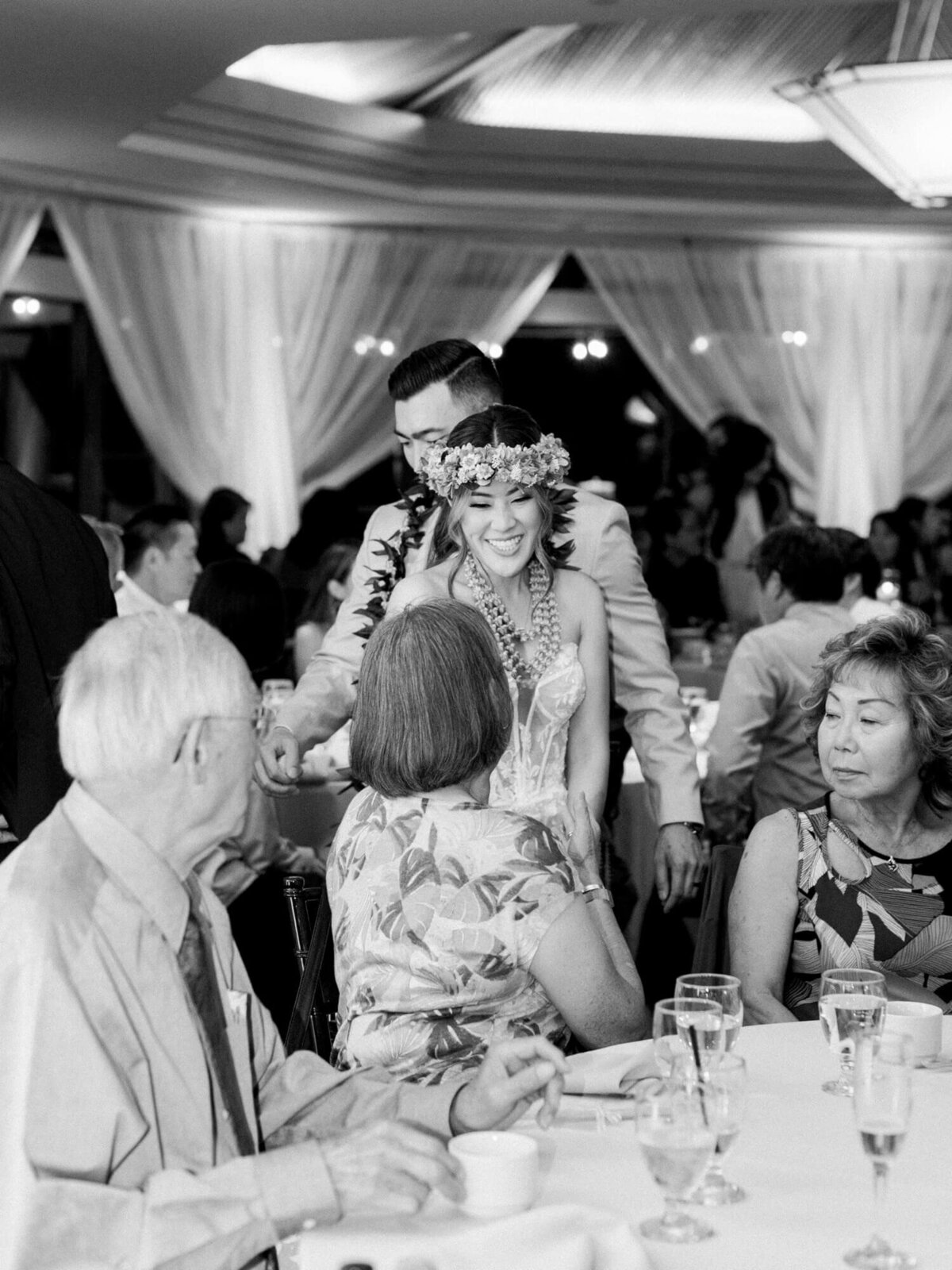 Black and white photo of a smiling bride in floral crown and groom at a wedding reception. Guests sit at a round table in a tent, creating a joyful atmosphere.