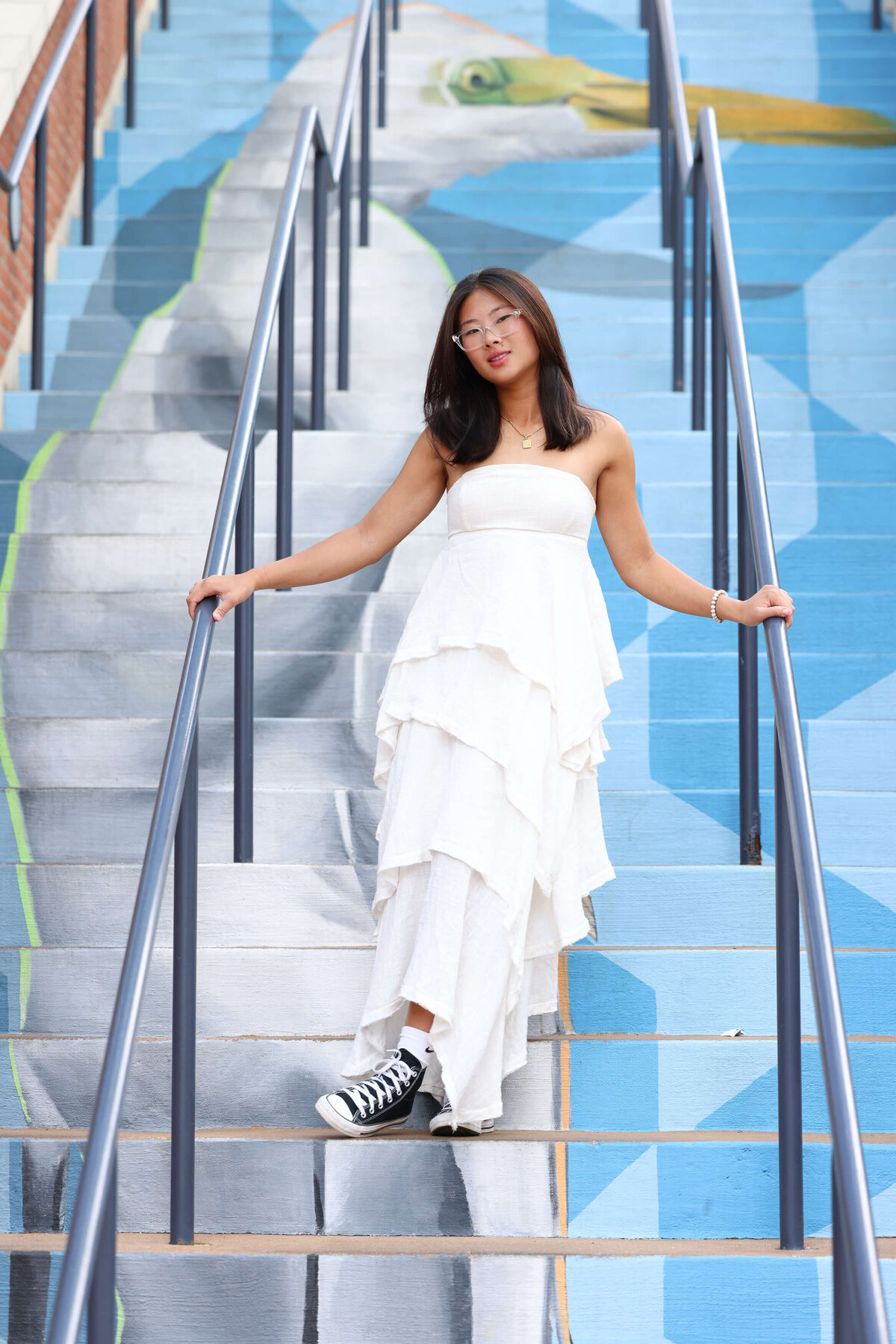 Annapolis High School Senior in flowing white dress and tennis shoes poses on blue heron mural stairs at Annapolis Town Center