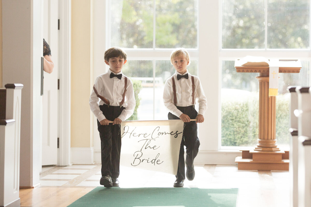 Two ring bearers hold a sign for the groom as they walk the isle and the ceremony begins at St. Franics at The Point in Point Clear, Alabama.