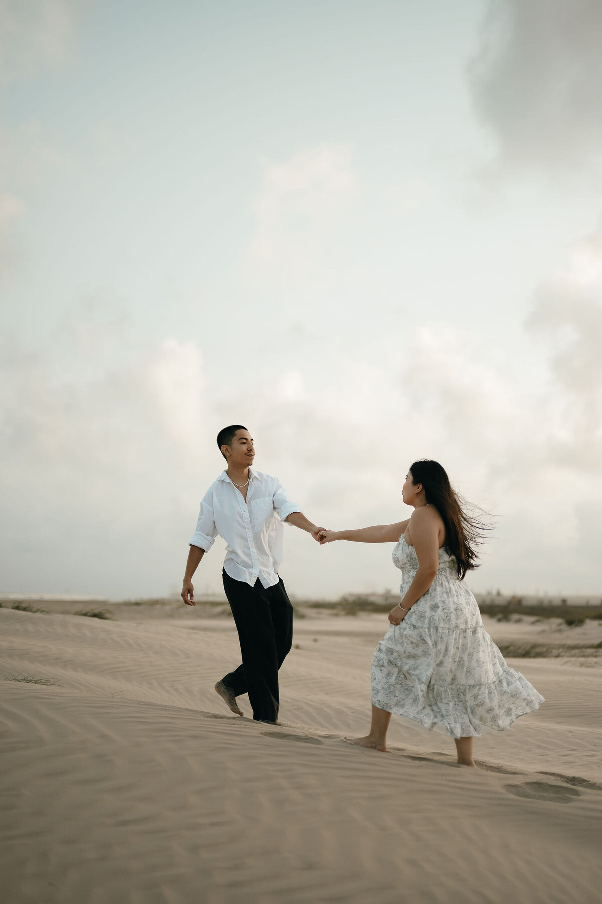 A couple joyfully runs hand-in-hand on a sandy beach, with the woman in a flowing dress and the man in a white shirt. A serene, cloudy sky is in the background.