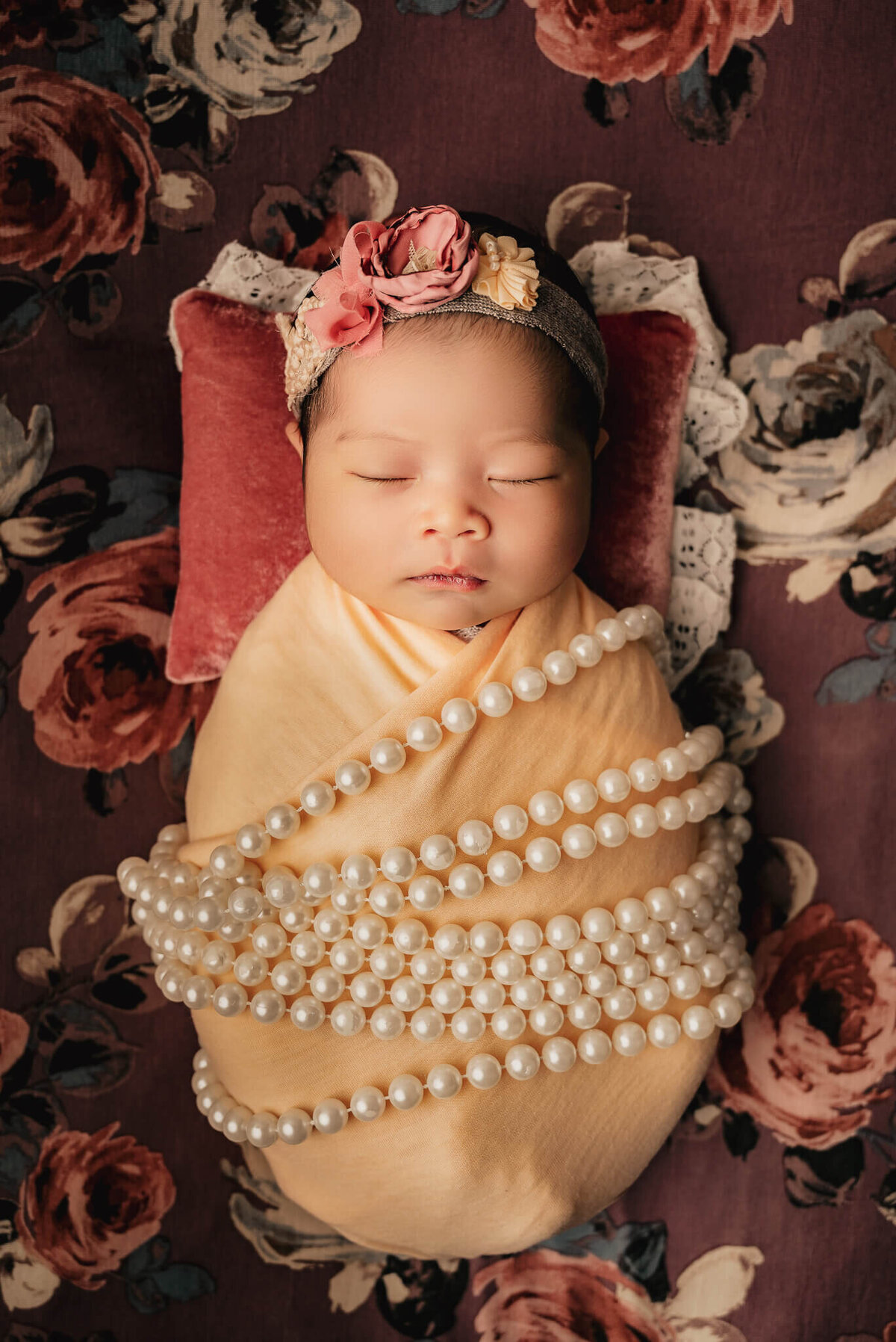 Infant girl wrapped in a peach wrap with white pearls sleeping on a rose patterned backdrop.