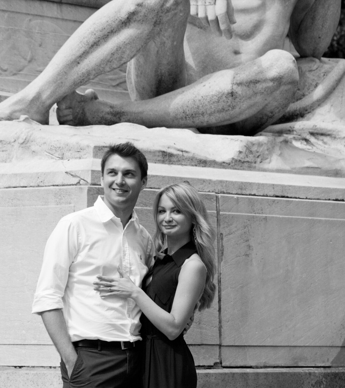 A black and white photograph of a smiling couple posed in front of the iconic statue at the main entrance of Central Park, New York. The man, clad  in white shirt and dark pants, gently holds the woman close, while she, adorned in a chick black dress, looks toward the camera with a soft smile. The grandeur of the statue, a symbol of the park's entrance, looms in the background, adding a timelss and artistic ambiance to the couple's portrait. -Photographed by Hans Gonzalez
