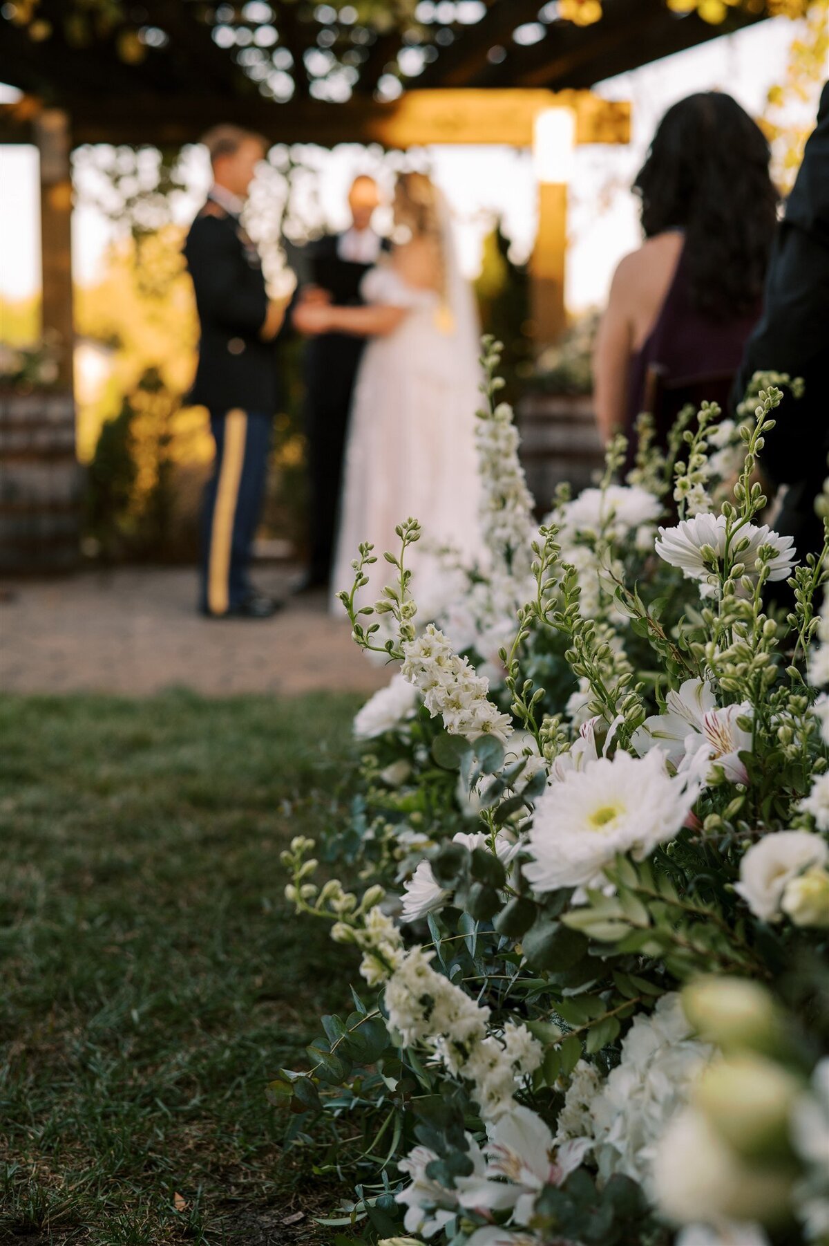 A wedding ceremony outdoors in Illinois with guests looking on, focusing on white flowers in the foreground with the bride and groom exchanging vows in the background.
