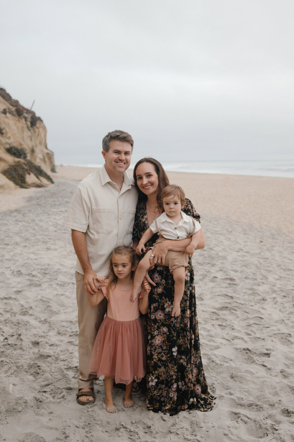 mom, dad, daughter and son stand on leucadia beach smiling at the camera with the cliffs and ocean in the background