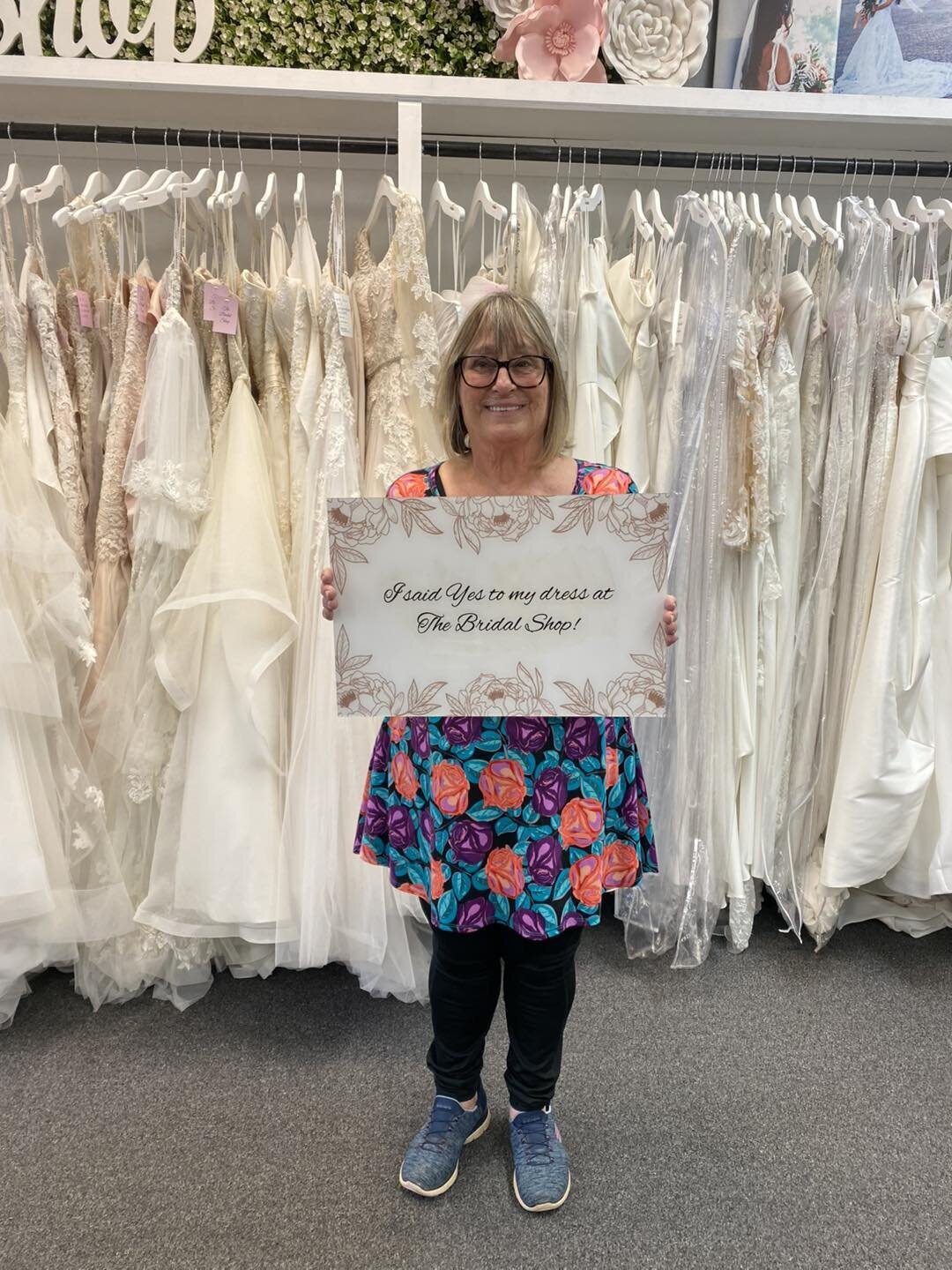 Woman standing in front of wedding dresses holding a sign announcing her dress choice