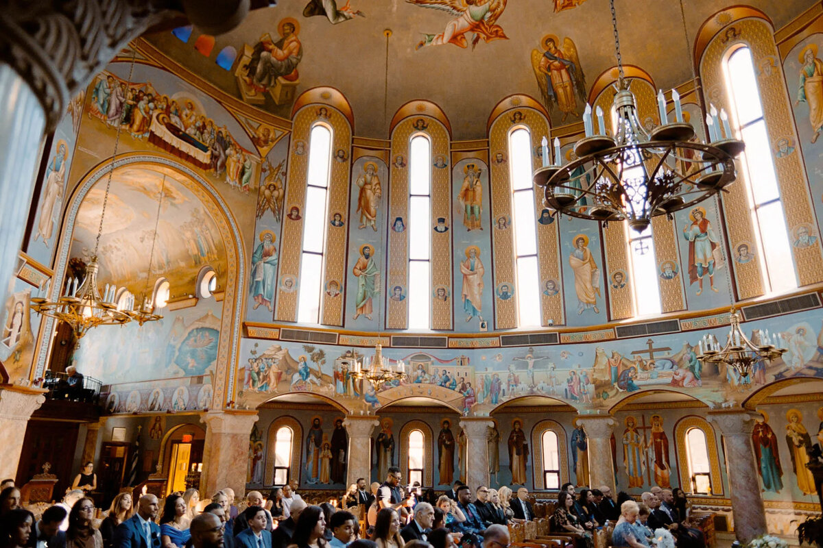 People seated inside a brightly lit church with ornate murals, stained glass windows, and elegant chandeliers—a setting a skilled NJ wedding photographer or film photographer NJ would beautifully capture amidst the arches and religious artwork.