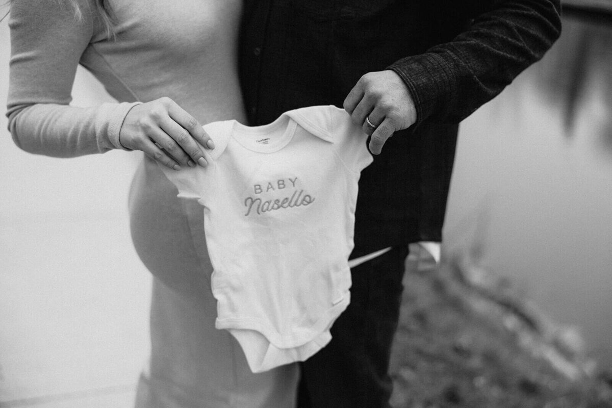 Man and woman holding baby onesie for baby announcement photos in Finger Lakes New York
