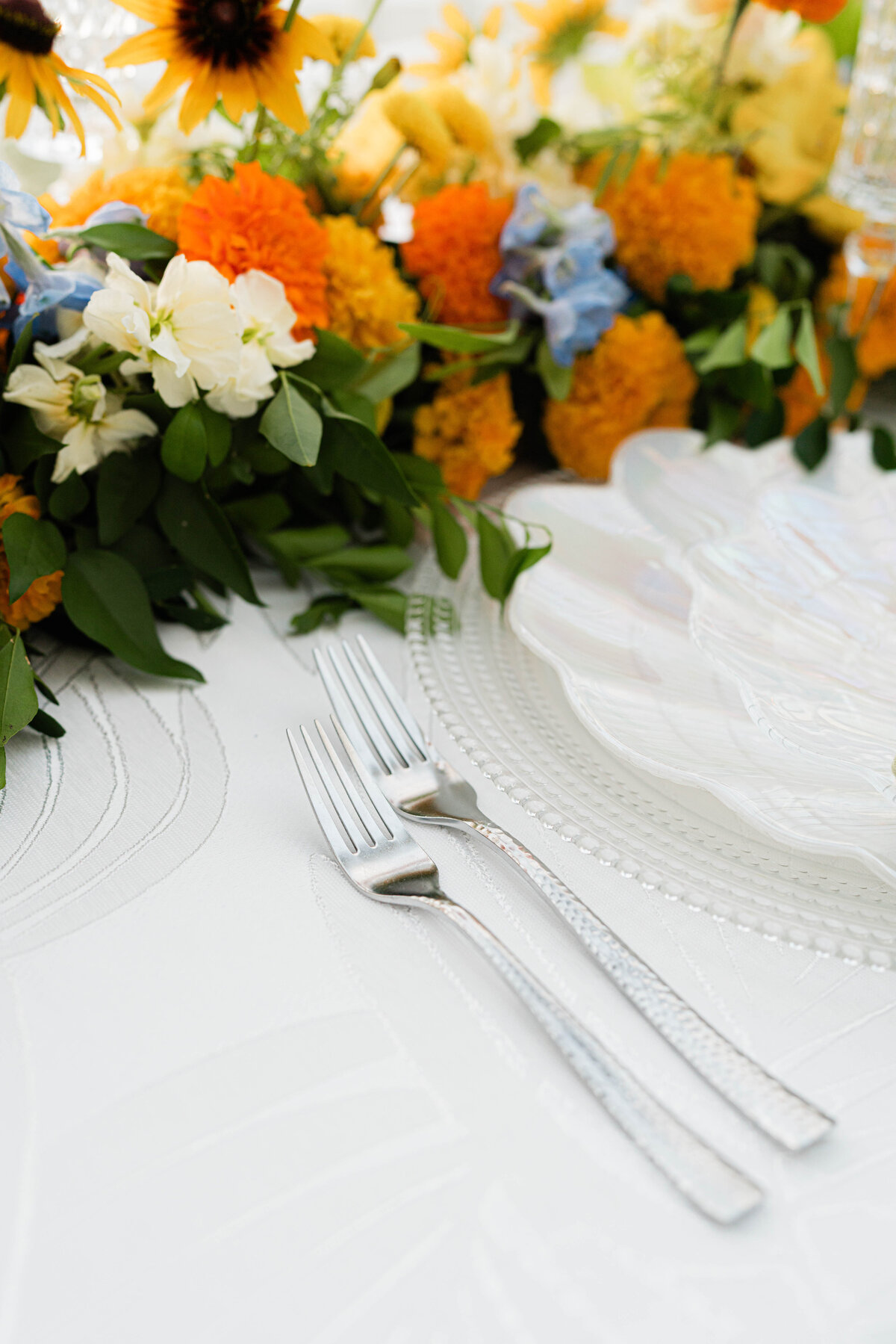 white on white palm leaf tablecloth with hammered silver forks and an iridescent seashell plate adorned with a white, orange, blue and green floral table runner