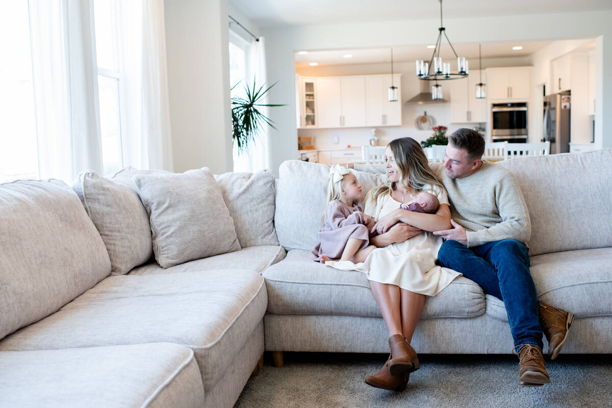 A mom and dad sit on a couch chatting with their toddler daughter while holding a sleeping newborn