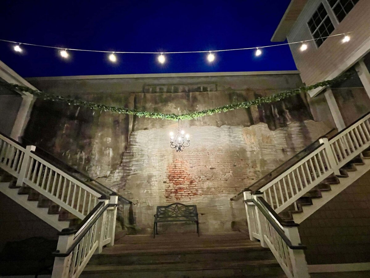 A nighttime scene of a stairway split in two, leading up to a brick wall with a bench beneath a hanging chandelier. String lights and greenery are draped above, with building exteriors on either side.