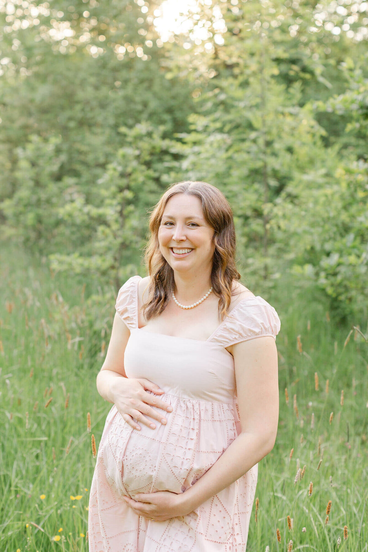 Beautiful pregnant mama in a pink dress with cap sleeves and some lace details and wearing a pearl necklace. She is holding her belly and smiling at the camera. She is standing in a field of tall golden grasses and green trees are behind her.