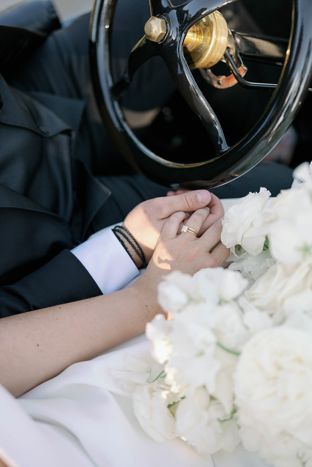 Close-up detail shot of bride and groom holding hands, wedding rings and bouquet visible, inside a vintage wedding car.