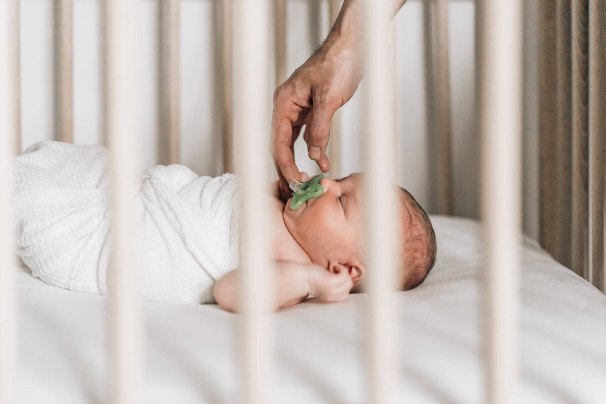 A through the crib slat photo of a partially swaddled newborn baby lying on her back, while her dad's hand reaches in from above to put in her pacifier.