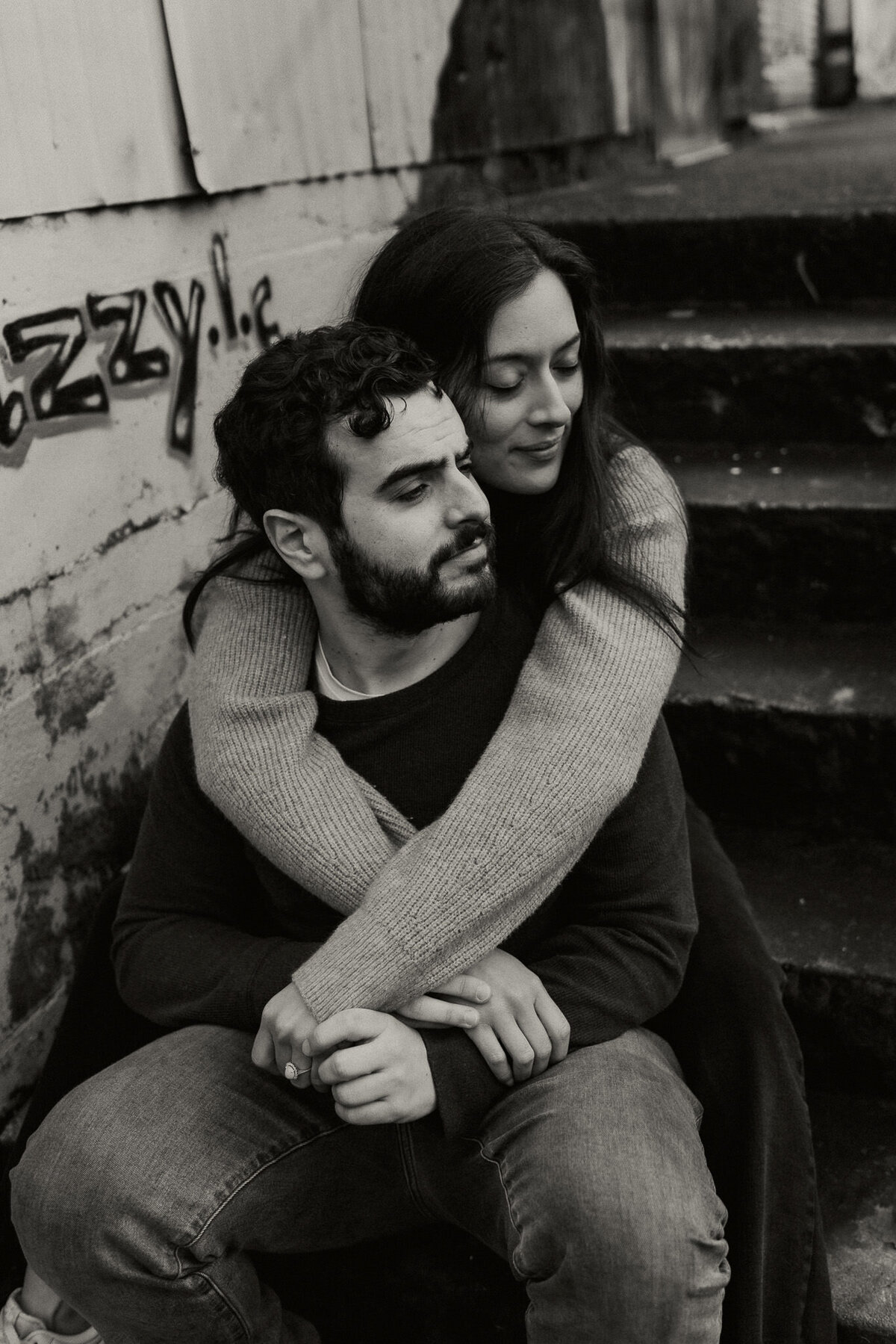 Urban engagement photo in black and white with couple sitting on steps with fiancee's arms wrapped around partner