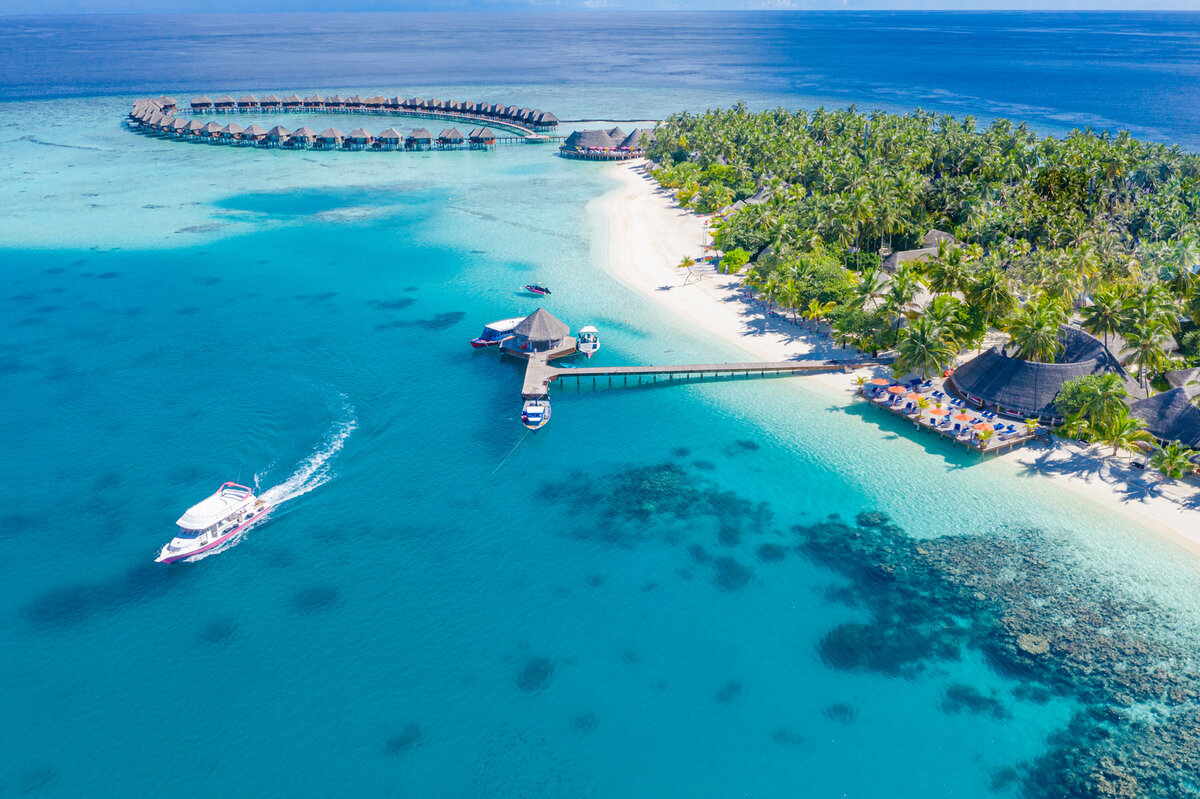 Aerial view of a tropical island resort with overwater bungalows arranged in a circular shape. A boat sails near a long pier extending from the sandy beach lined with palm trees. The surrounding ocean is a vibrant turquoise color.